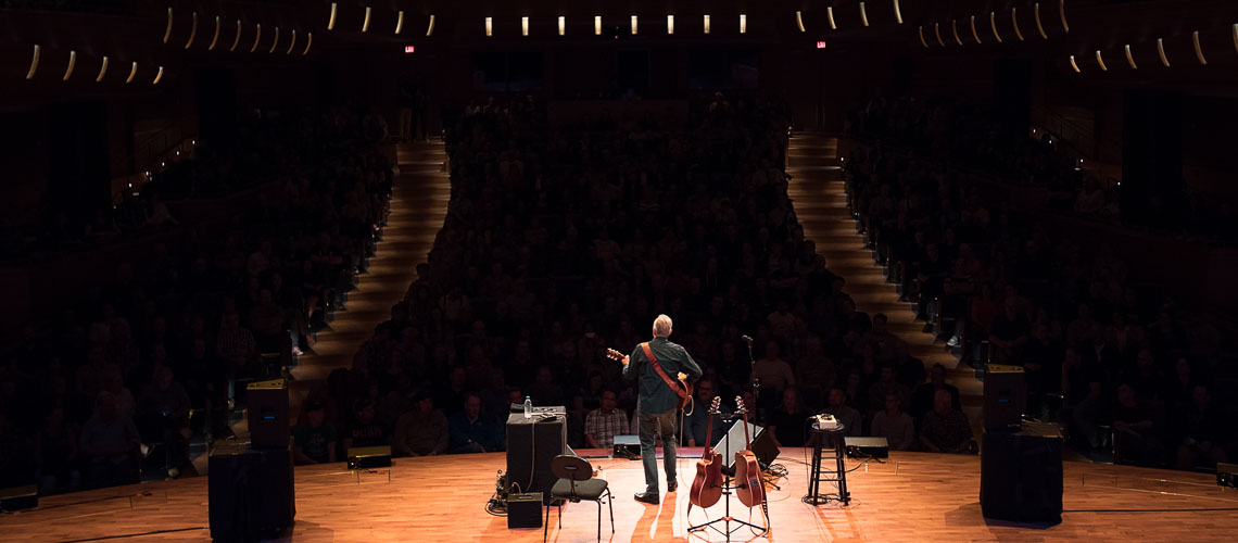A man, viewed from behind, playing the guitar on the stage of the Bella Concert Hall